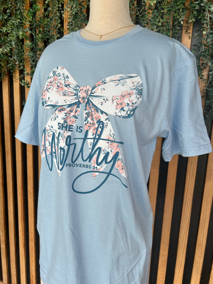 Close-up of the She Is Worthy Floral Bow Graphic Tee in light blue featuring a large floral bow graphic with the inspirational "She Is Worthy" Proverbs 31:25 design. Displayed on a mannequin with a crew neckline and short sleeves.