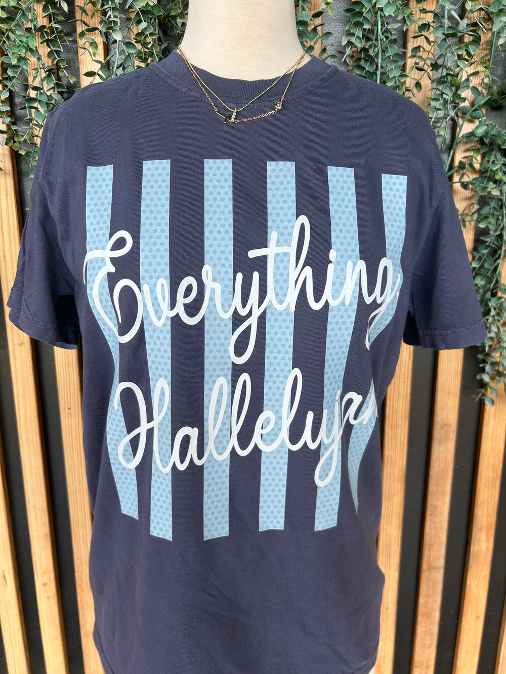 Close-up of navy faith graphic tee featuring Everything Hallelujah script over light blue striped background.