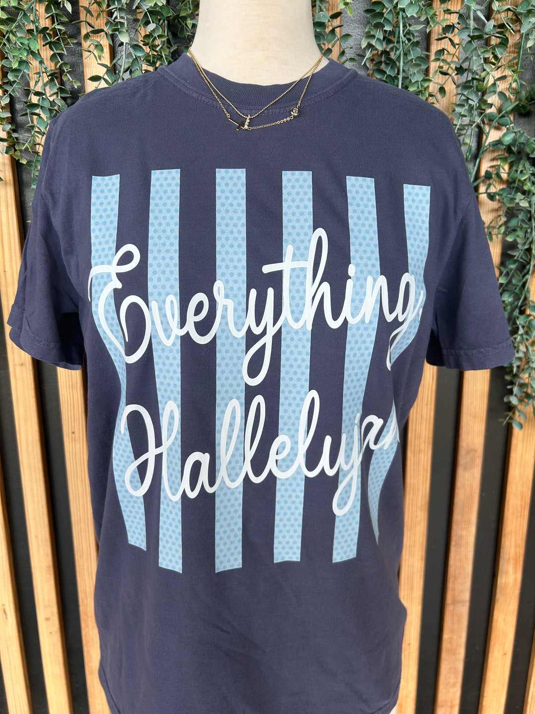 Close-up of navy faith graphic tee featuring Everything Hallelujah script over light blue striped background.