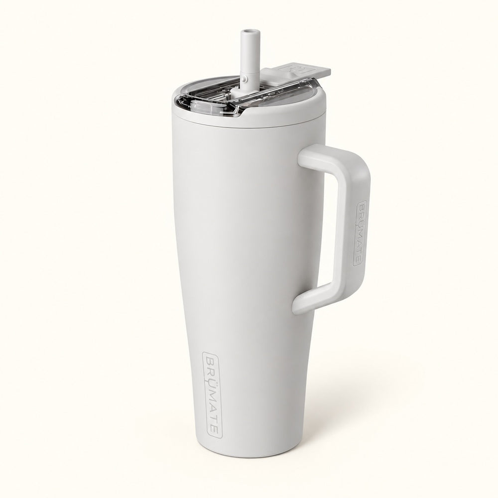 BrüMate Era Flip 40oz insulated straw tumbler in Dune featuring a soft matte light grey-beige finish, large coordinating carrying handle, covered flip-down straw lid shown open, and vertical BrüMate branding. The tall 40oz leakproof water tumbler features a cupholder-friendly base and is displayed against a soft cream studio background.