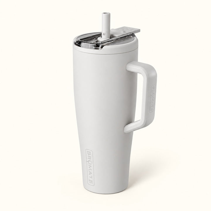 BrüMate Era Flip 40oz insulated straw tumbler in Dune featuring a soft matte light grey-beige finish, large coordinating carrying handle, covered flip-down straw lid shown open, and vertical BrüMate branding. The tall 40oz leakproof water tumbler features a cupholder-friendly base and is displayed against a soft cream studio background.