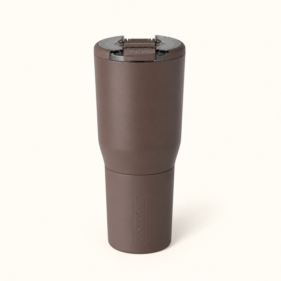 BrüMate Nav 35oz insulated travel tumbler in Java featuring a matte brown finish, leakproof BevLock lid in the closed position, tall cylindrical body, and removable color-matched protective sleeve with embossed BrüMate branding. Displayed upright against a soft cream studio background.