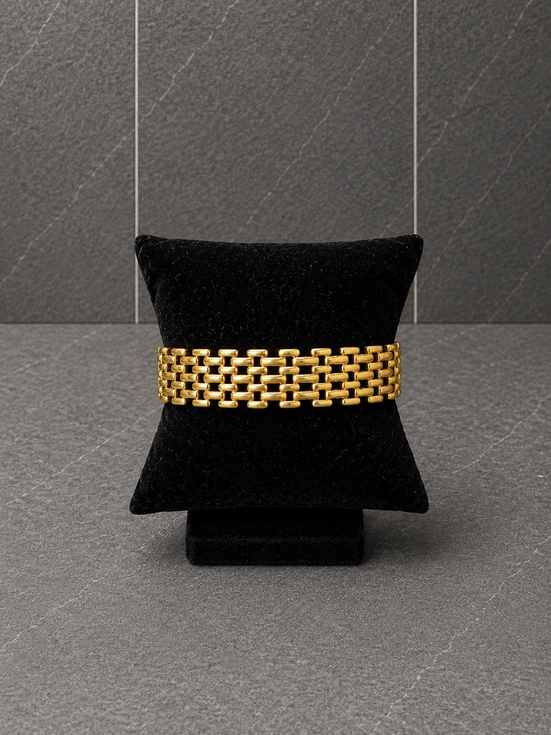 A gold-toned woven link bracelet displayed on a black velvet jewelry pillow against a dark gray textured background.