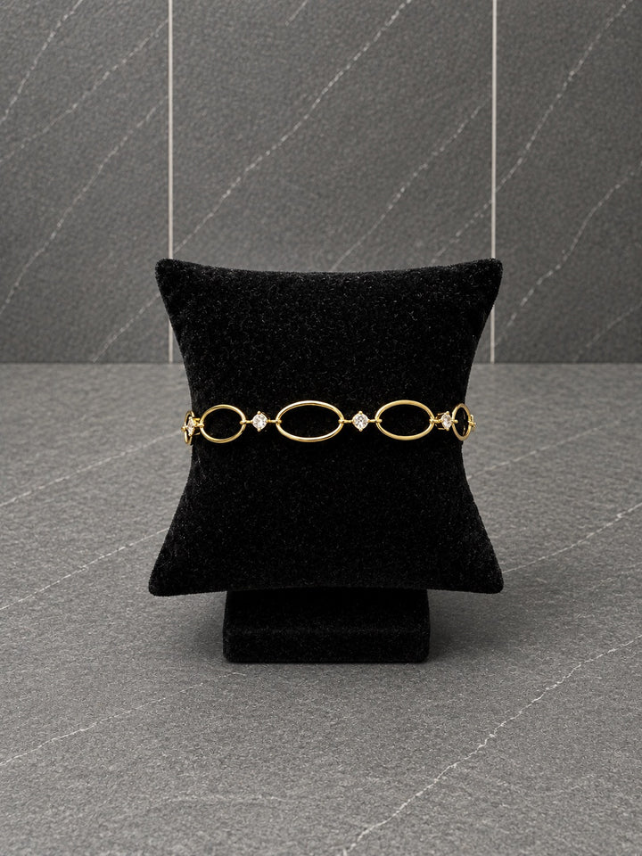 A gold chain bracelet with oval links and small clear stones, displayed on a black velvet jewelry cushion against a gray textured background.