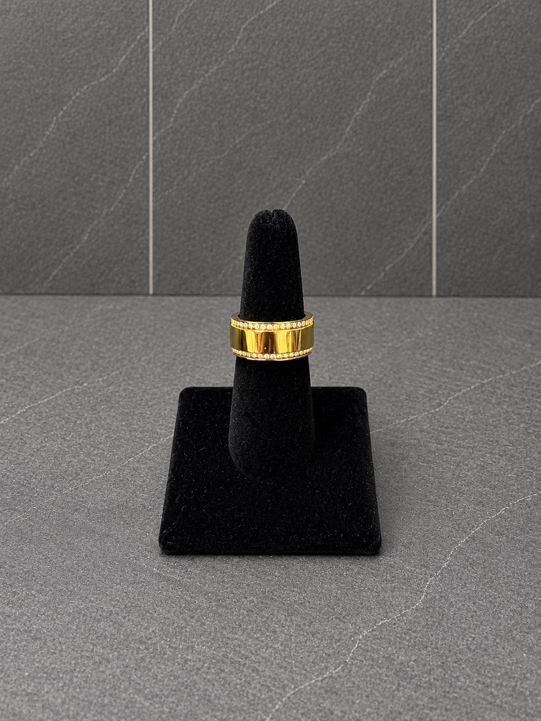 A gold band ring  with crystal accents displayed on a black ring stand against a dark gray textured background.