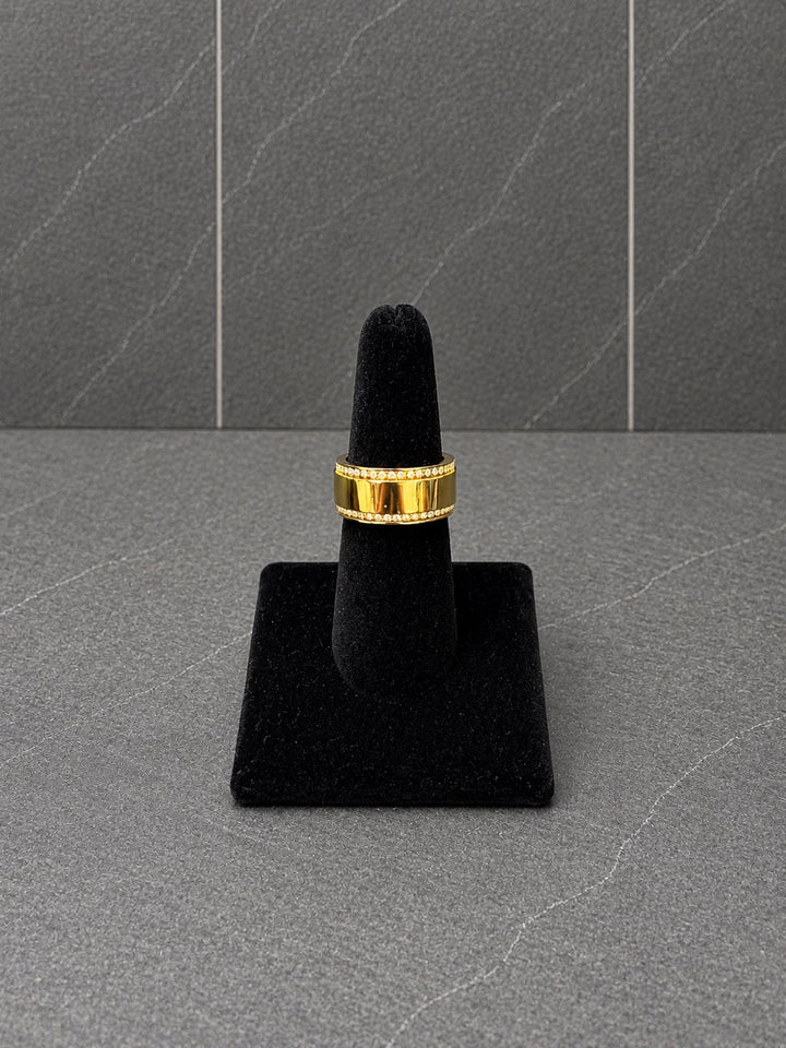 A gold band ring  with crystal accents displayed on a black ring stand against a dark gray textured background.