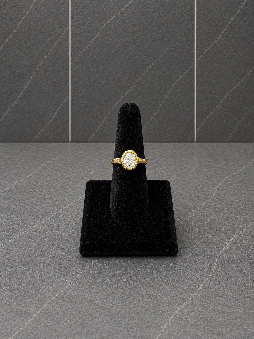 A yellow gold ring with a round-cut clear gemstone displayed on a black velvet ring stand against a dark gray textured background.