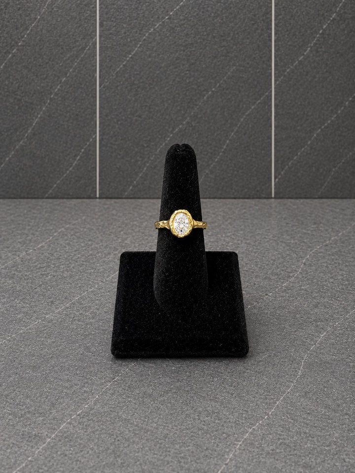 A yellow gold ring with a round-cut clear gemstone displayed on a black velvet ring stand against a dark gray textured background.