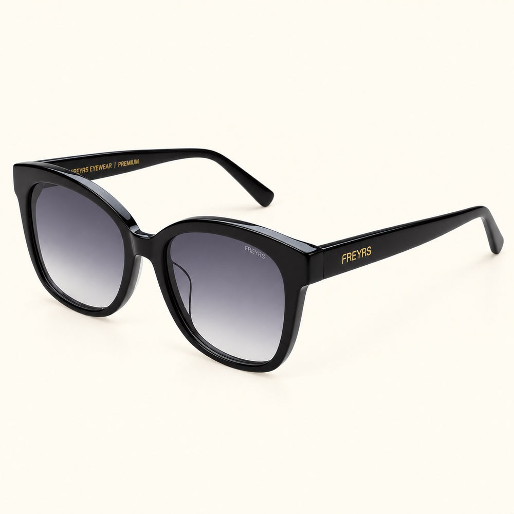 Angled view of FREYRS Amber Black cat eye sunglasses showing thick acetate frames, gray gradient lenses, wide black temples, and gold FREYRS branding.