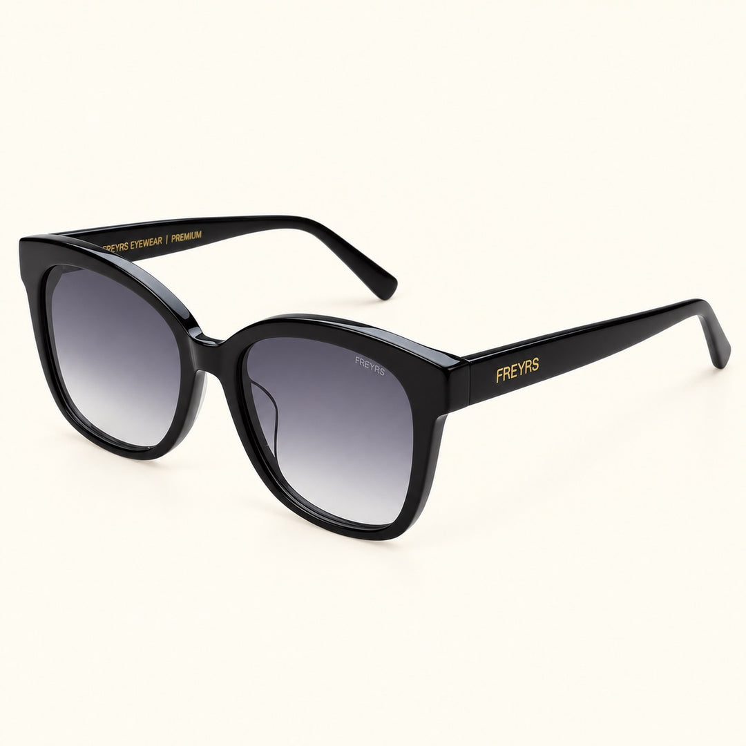 Angled view of FREYRS Amber Black cat eye sunglasses showing thick acetate frames, gray gradient lenses, wide black temples, and gold FREYRS branding.