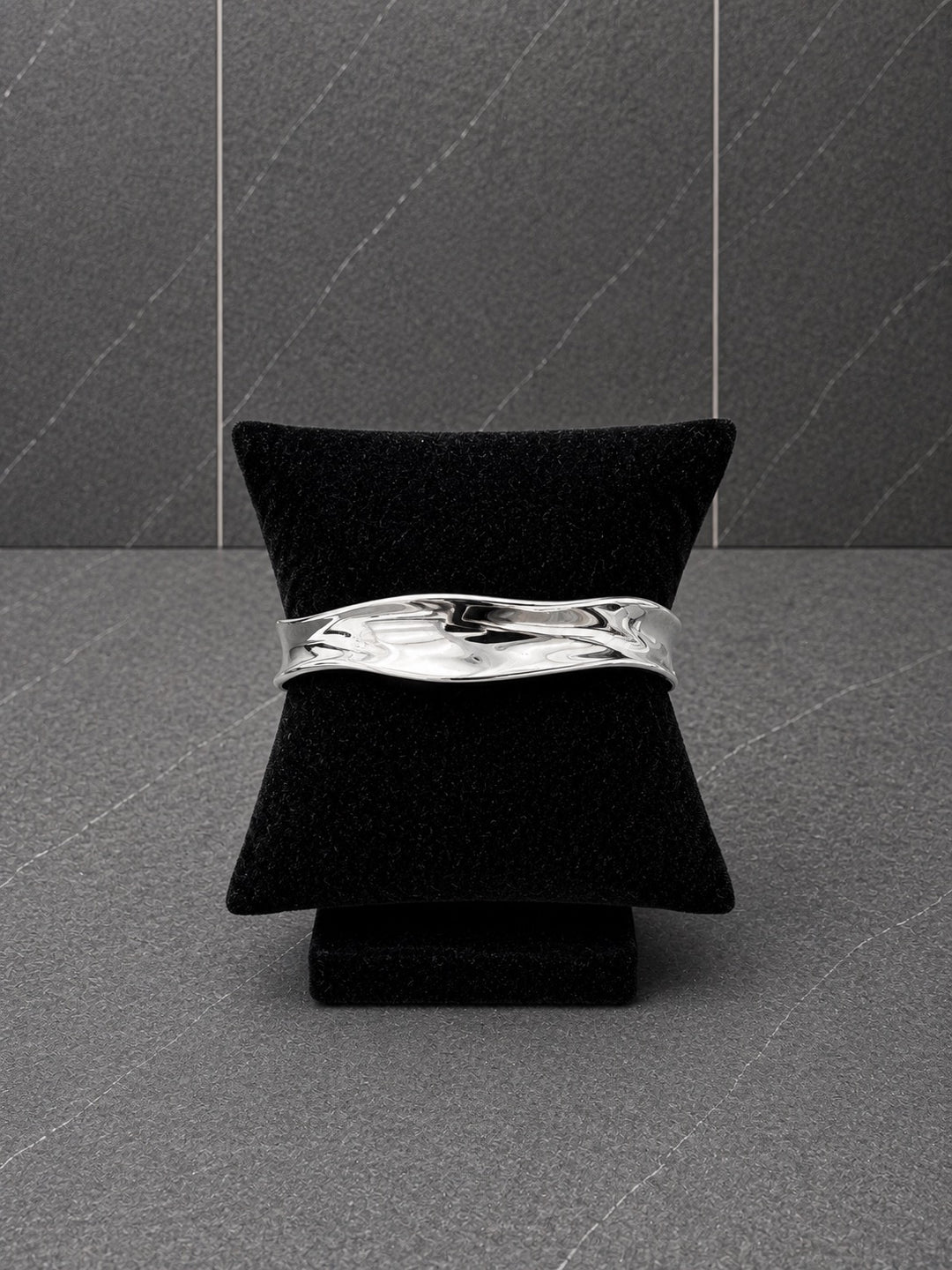 A polished silver wavy cuff bracelet displayed on a black velvet jewelry stand against a dark gray textured background.
