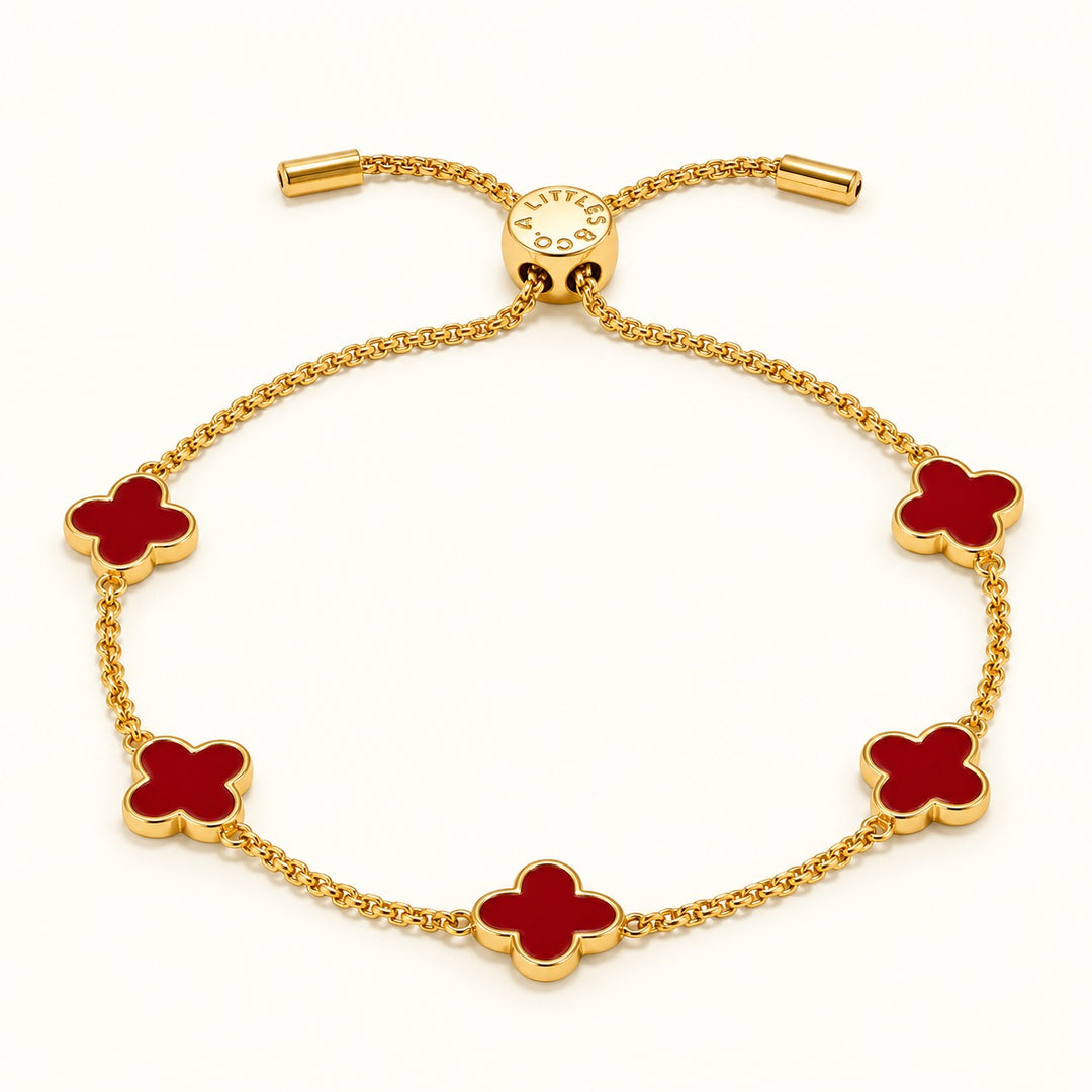 A Littles &amp; Co. gold-tone adjustable bracelet featuring five red enamel clover charms, a sliding bolo clasp, and gold chain ends, arranged in an oval on a soft cream studio background.