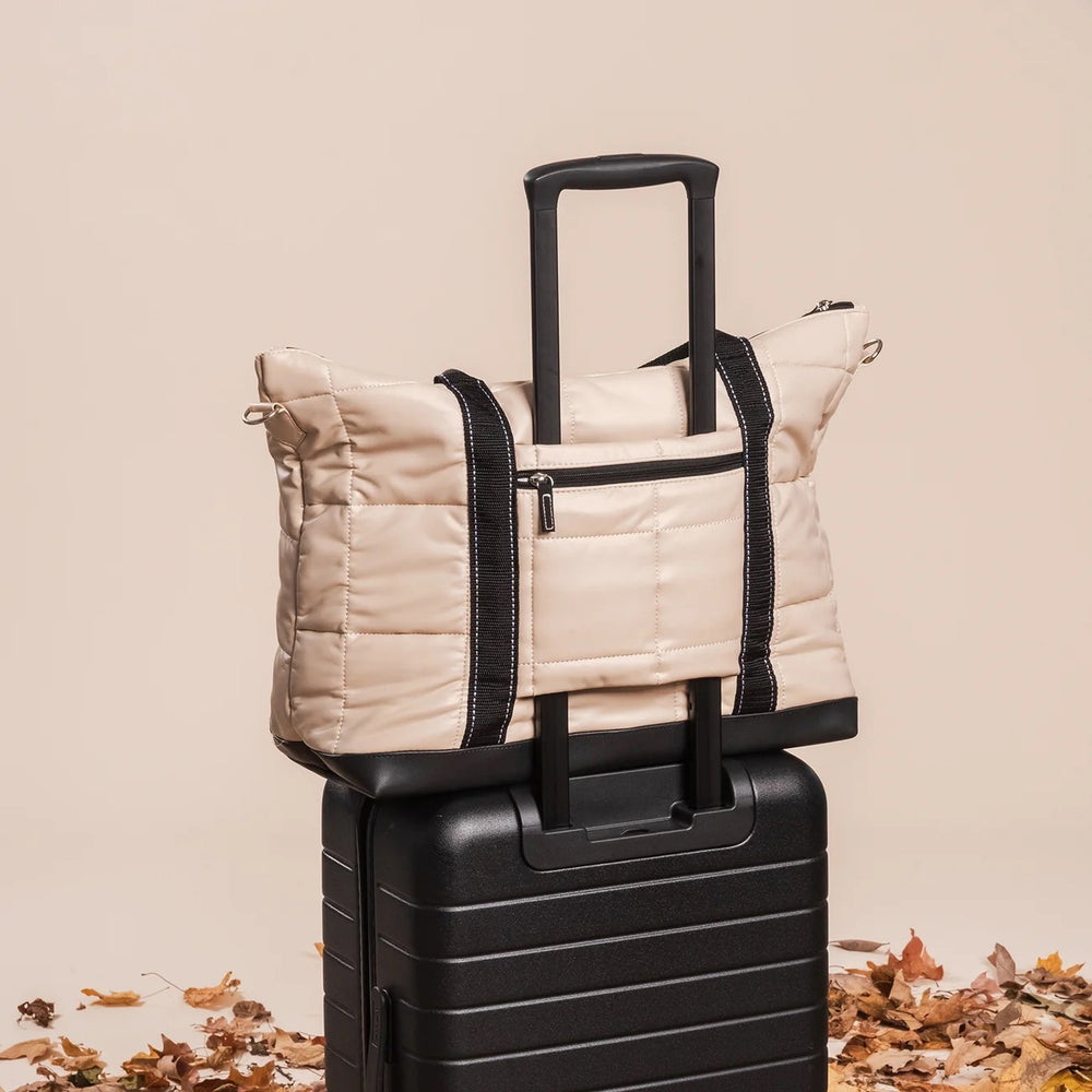 A beige quilted puffer tote bag with black trim and handles is draped over the handle of a black hard-shell suitcase, set against a warm beige background with scattered autumn leaves at the base.