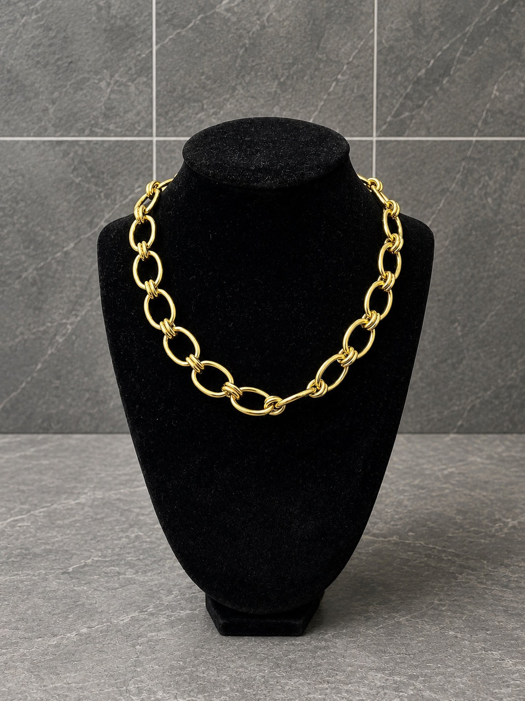 A chunky gold chain necklace with oversized interlocking oval links, displayed on a black velvet jewelry bust against a gray tiled background.
