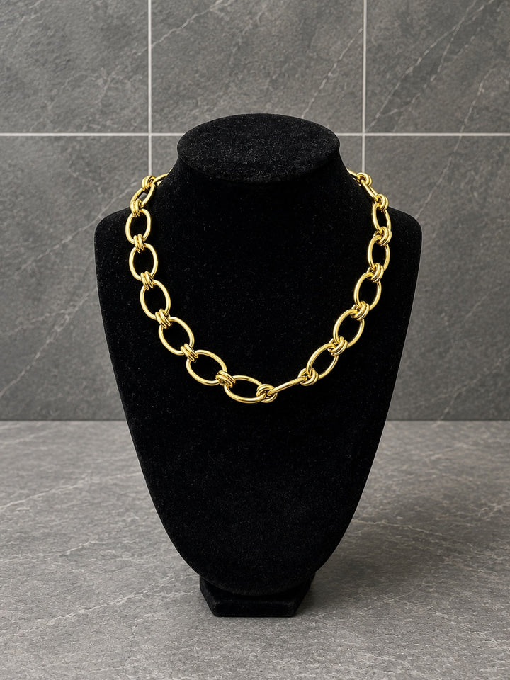 A chunky gold chain necklace with oversized interlocking oval links, displayed on a black velvet jewelry bust against a gray tiled background.