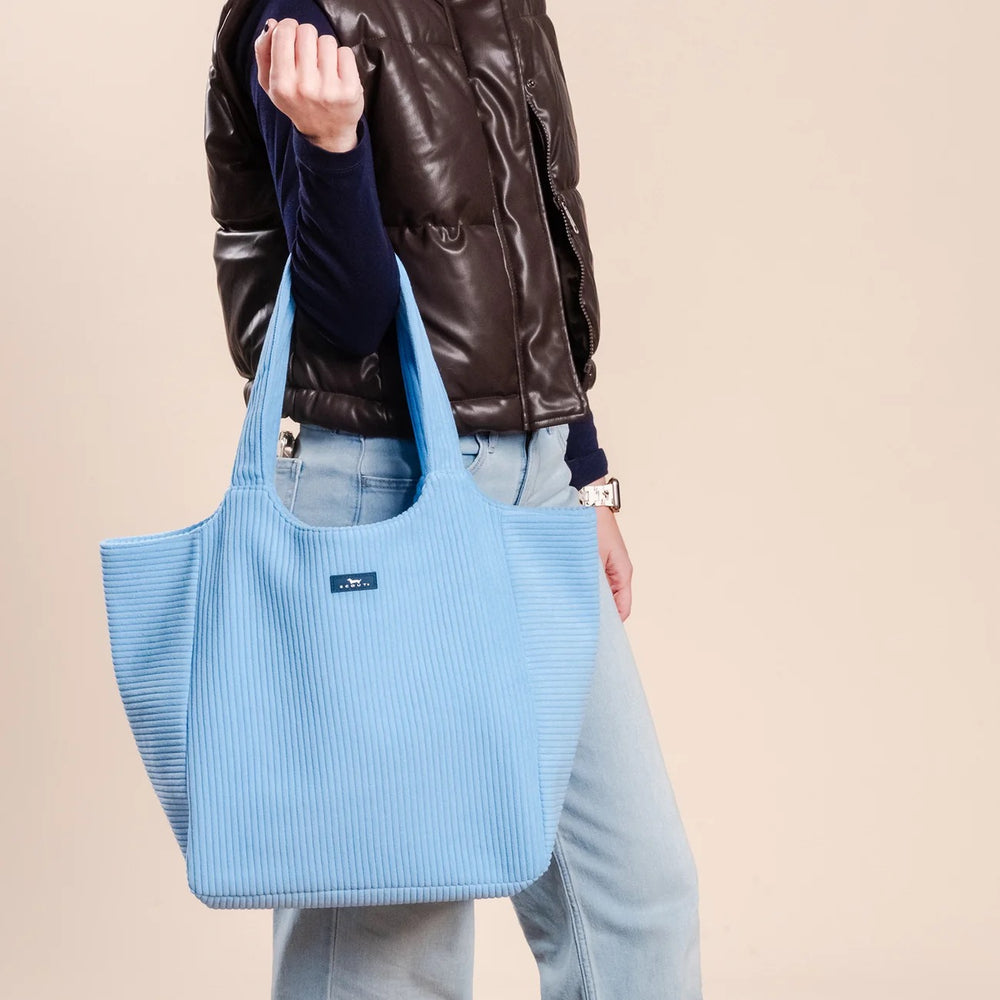 A person wearing a dark brown puffer jacket and light blue jeans carries a large light blue ribbed fabric tote bag with double shoulder straps against a plain beige background.
