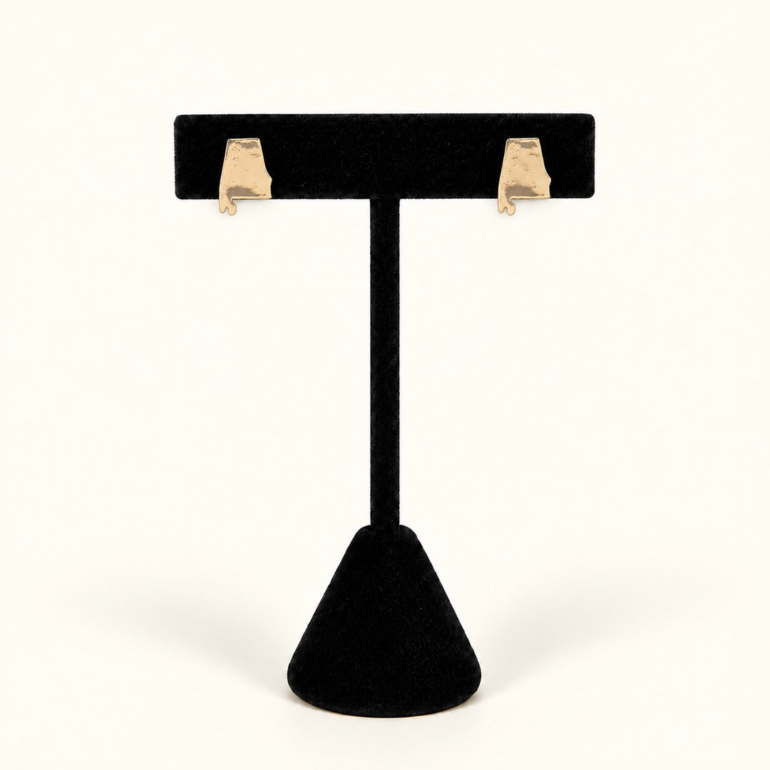 A pair of small gold Alabama state stud earrings displayed on a black velvet T-stand against a clean off-white background.