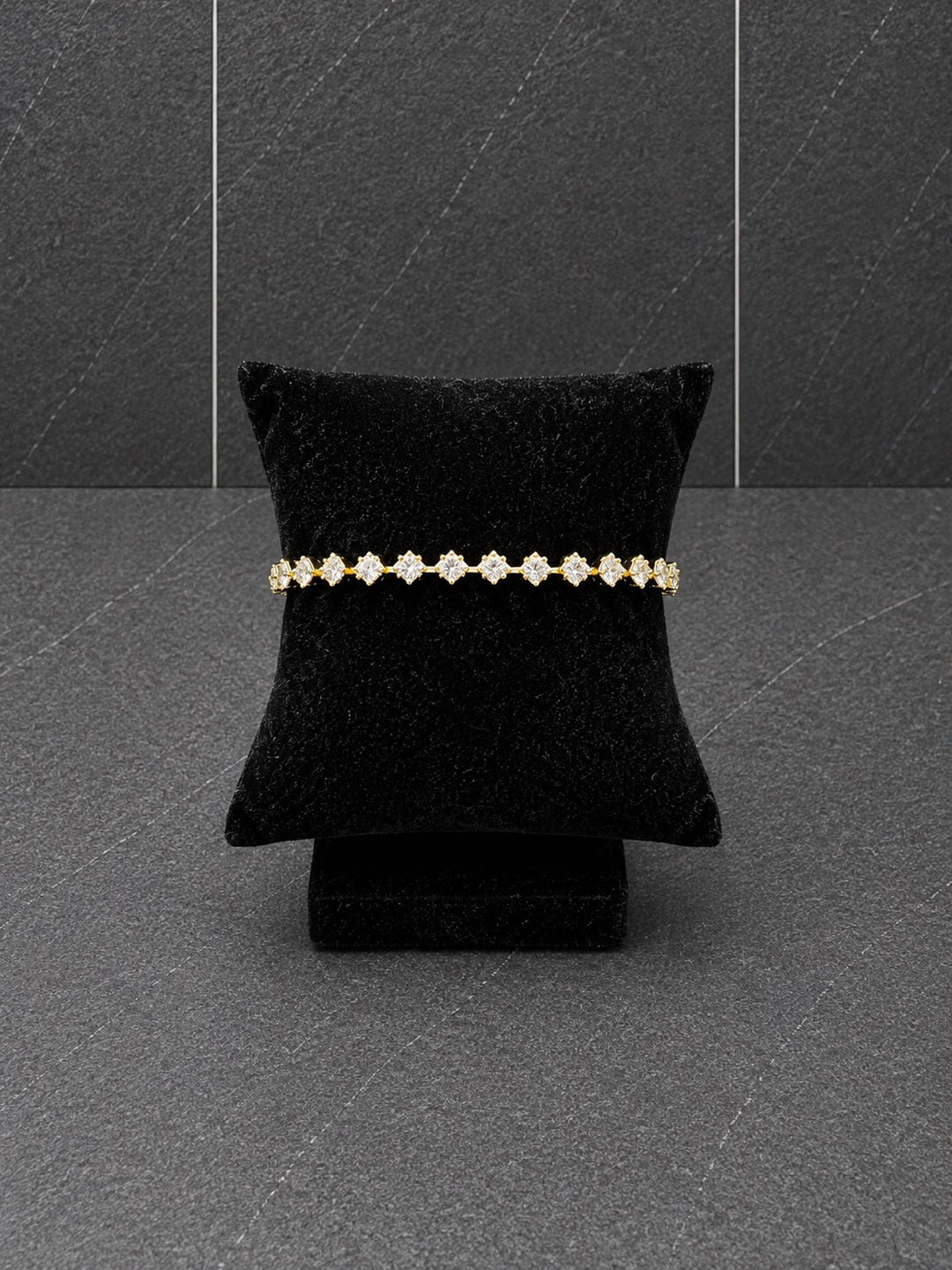 A delicate gold bracelet with small clear floral inspired gemstones displayed on a black velvet jewelry pillow against a dark gray textured background.