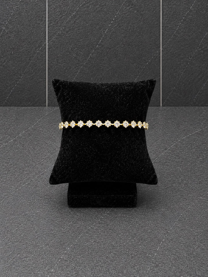 A delicate gold bracelet with small clear floral inspired gemstones displayed on a black velvet jewelry pillow against a dark gray textured background.