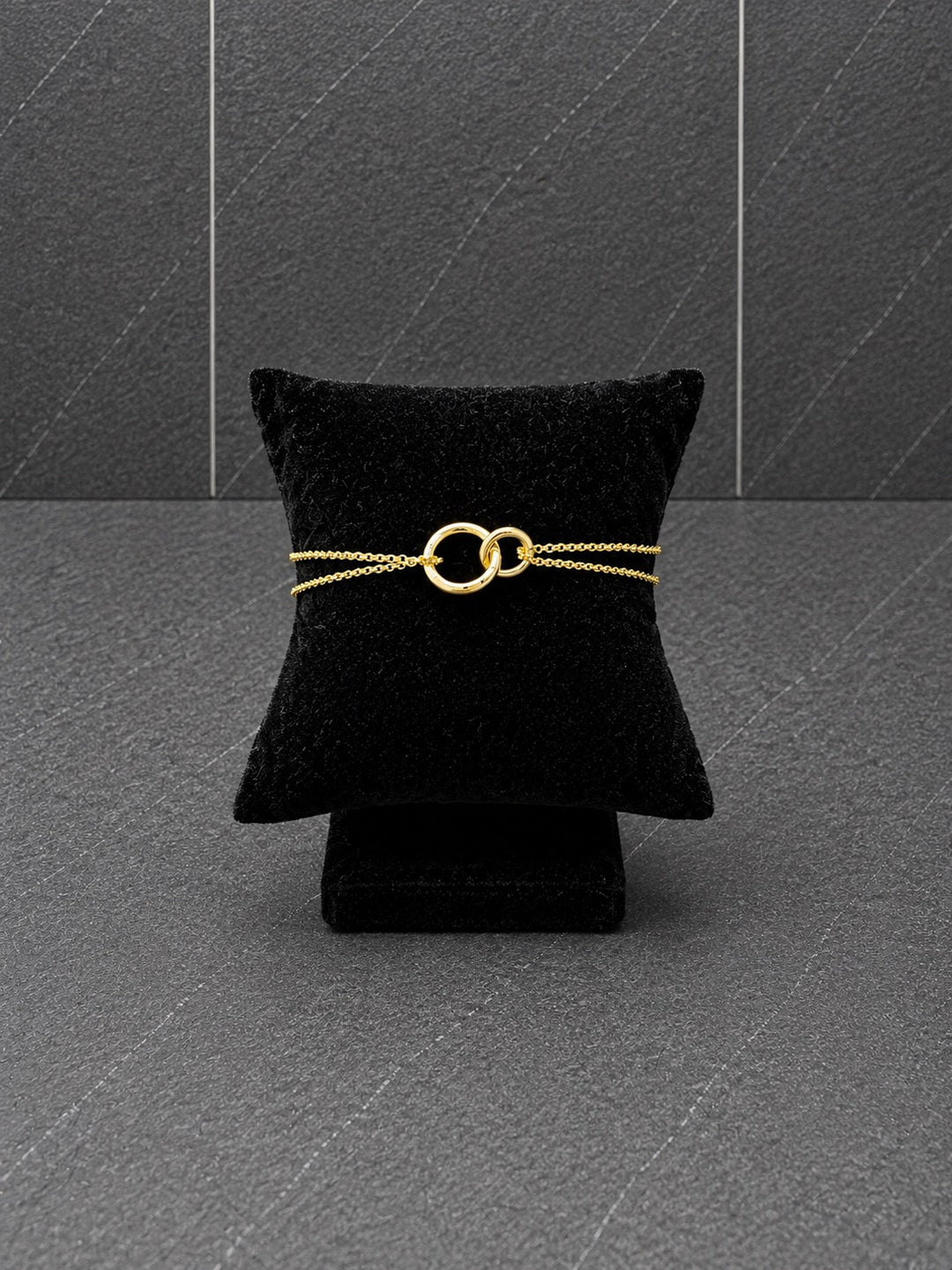 A delicate gold chain bracelet with two interlocking rings, displayed on a black velvet jewelry pillow against a dark gray textured background.