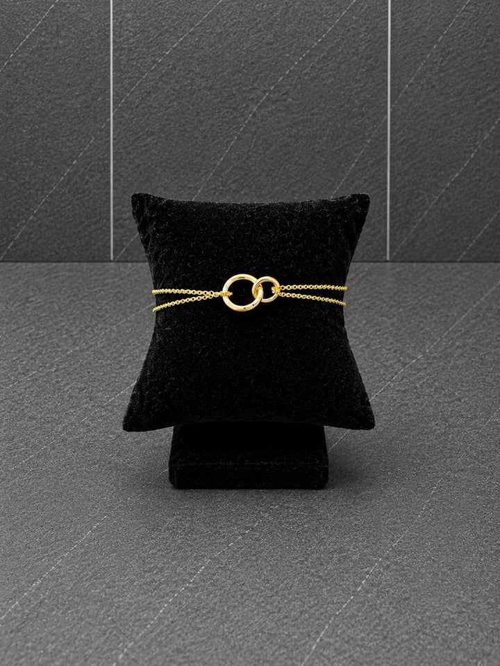 A delicate gold chain bracelet with two interlocking rings, displayed on a black velvet jewelry pillow against a dark gray textured background.