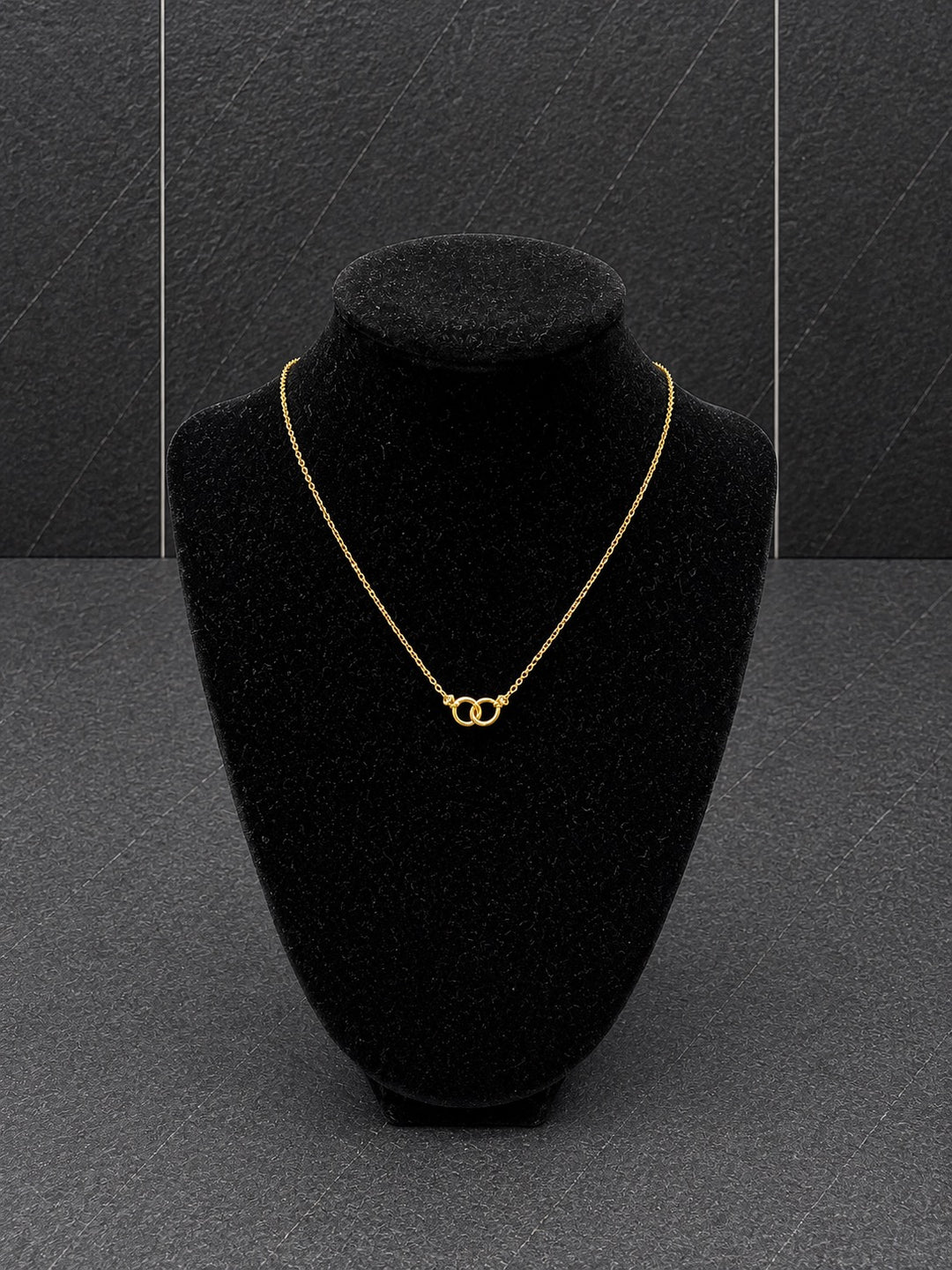 A delicate gold chain necklace with a double interlocking ring pendant, displayed on a black velvet jewelry bust against a dark gray textured background.