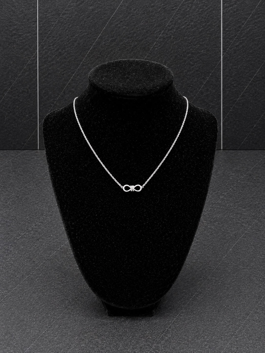 A delicate silver infinity necklace displayed on a black velvet jewelry bust against a dark gray textured background.