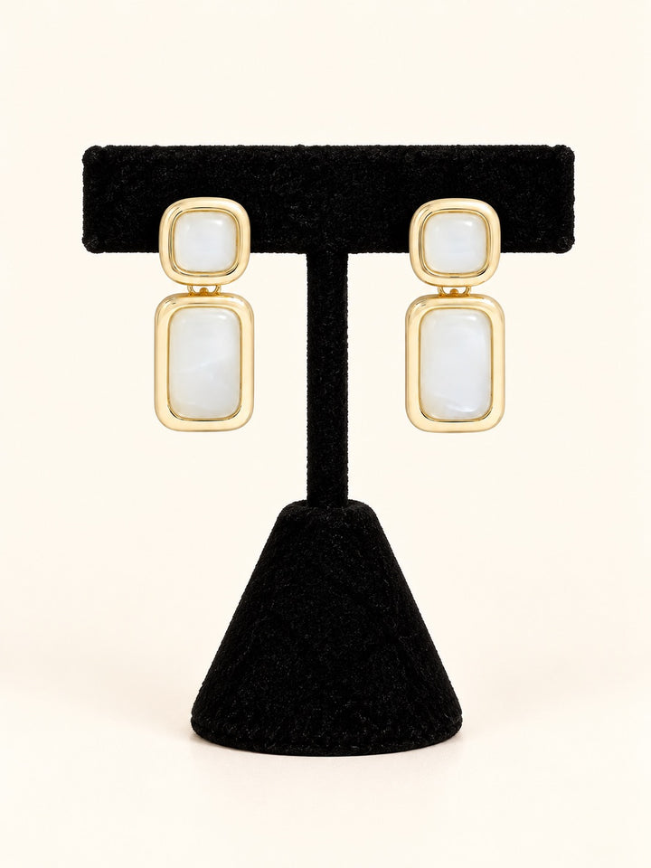 Laurel Earrings in Cream featuring glossy ivory-white resin accents, polished gold-tone frames, and geometric drop design on a black velvet stand.