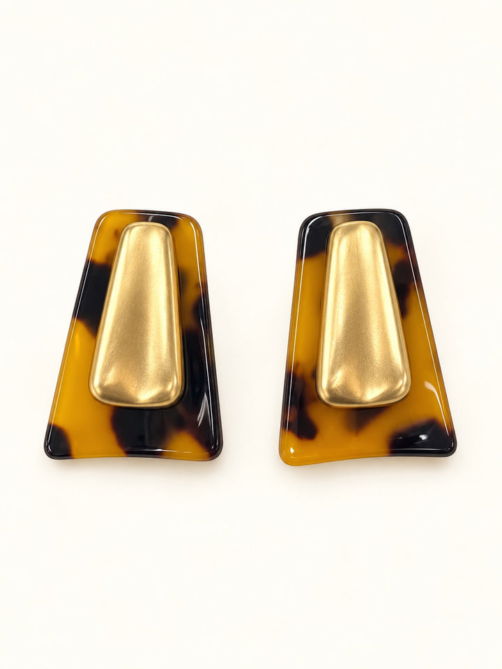 Close-up of Michelle McDowell Willa Tortoise Shell Earrings featuring glossy amber and dark brown geometric frames with brushed gold-tone center details on a cream studio background.