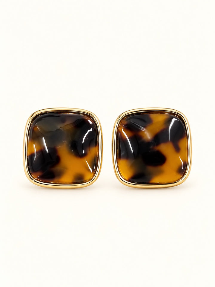 Close-up of Ruth Tortoise Earrings showing rounded square studs with rich amber, caramel, and black tortoiseshell patterns surrounded by polished gold-tone frames on a cream studio background.