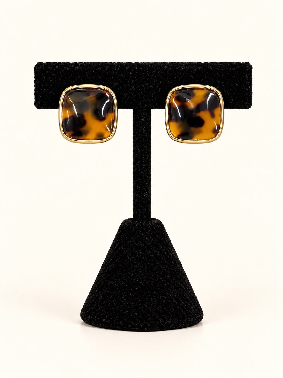 Ruth Tortoise Square Stud Earrings featuring glossy amber and dark brown marbled centers framed in polished gold-tone borders, displayed on a black velvet earring stand against a cream background.