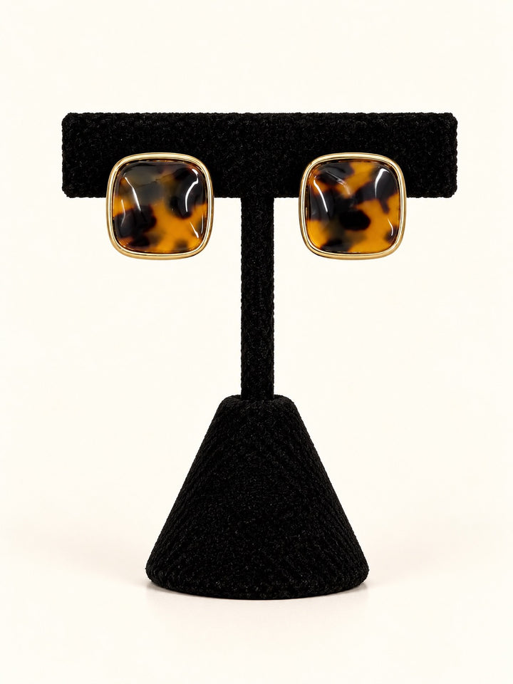 Ruth Tortoise Square Stud Earrings featuring glossy amber and dark brown marbled centers framed in polished gold-tone borders, displayed on a black velvet earring stand against a cream background.