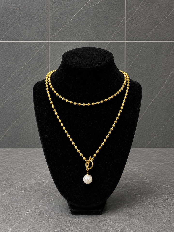 A double-strand gold necklace with a small white pearl pendant, displayed on a black velvet jewelry bust against a dark gray tiled background.