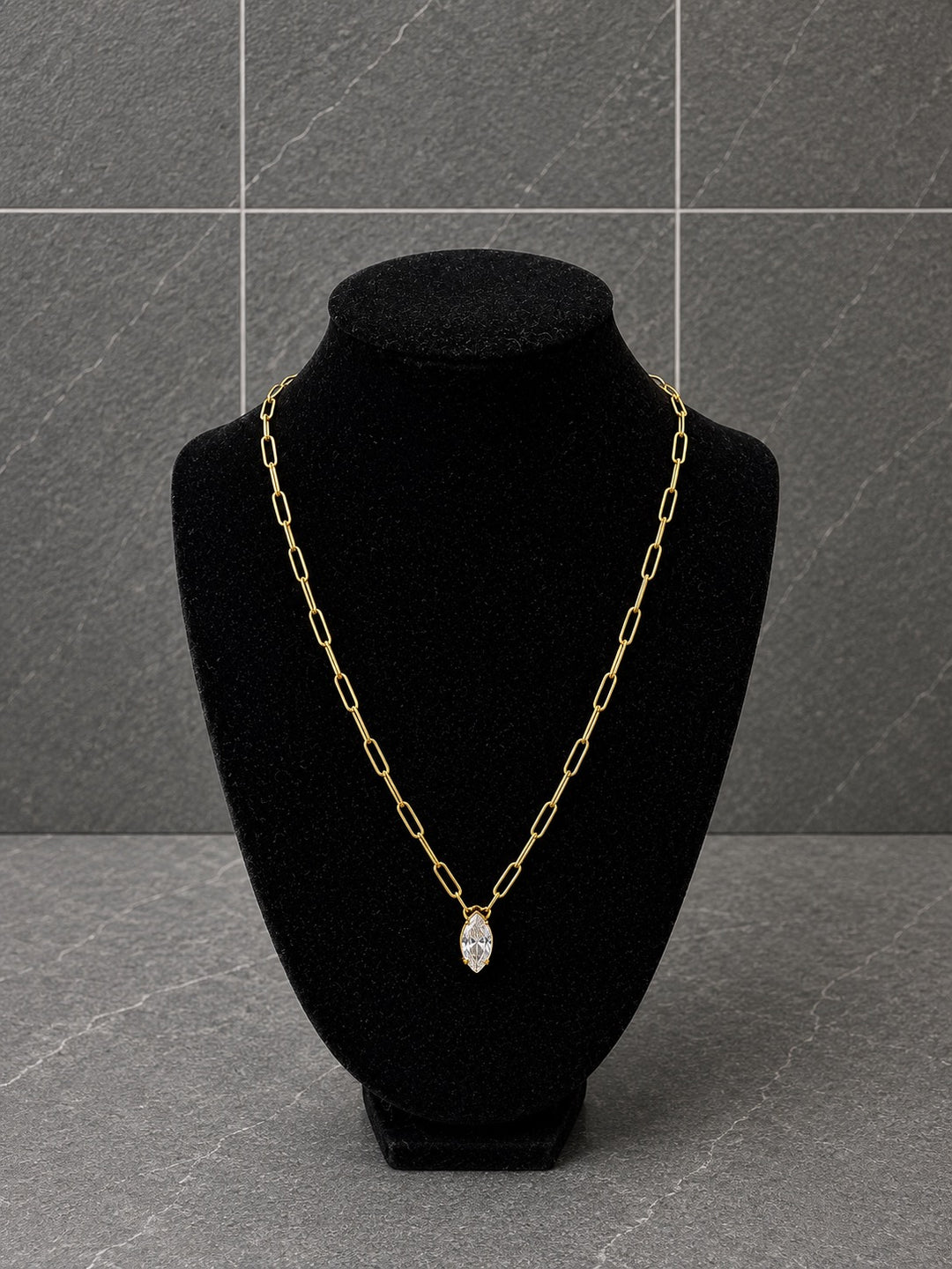 A gold chain necklace with a marquise-shaped clear gemstone pendant, displayed on a black velvet jewelry bust against a gray tiled background.