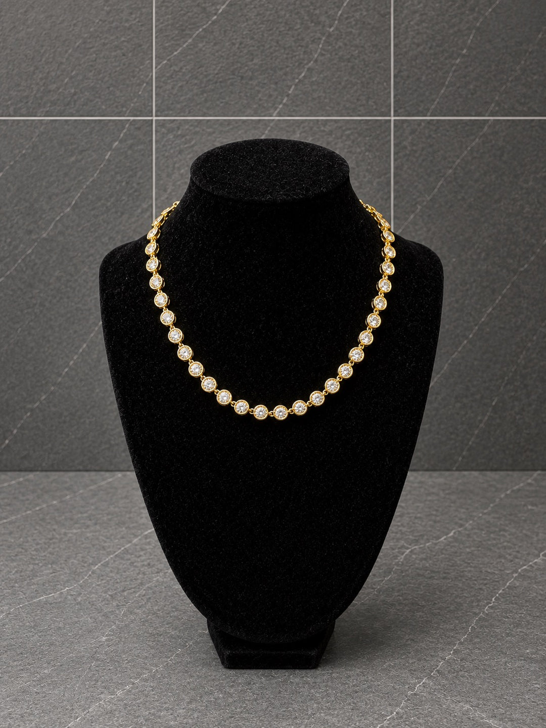 A gold-toned beaded necklace displayed on a black velvet jewelry bust against a gray tiled background.