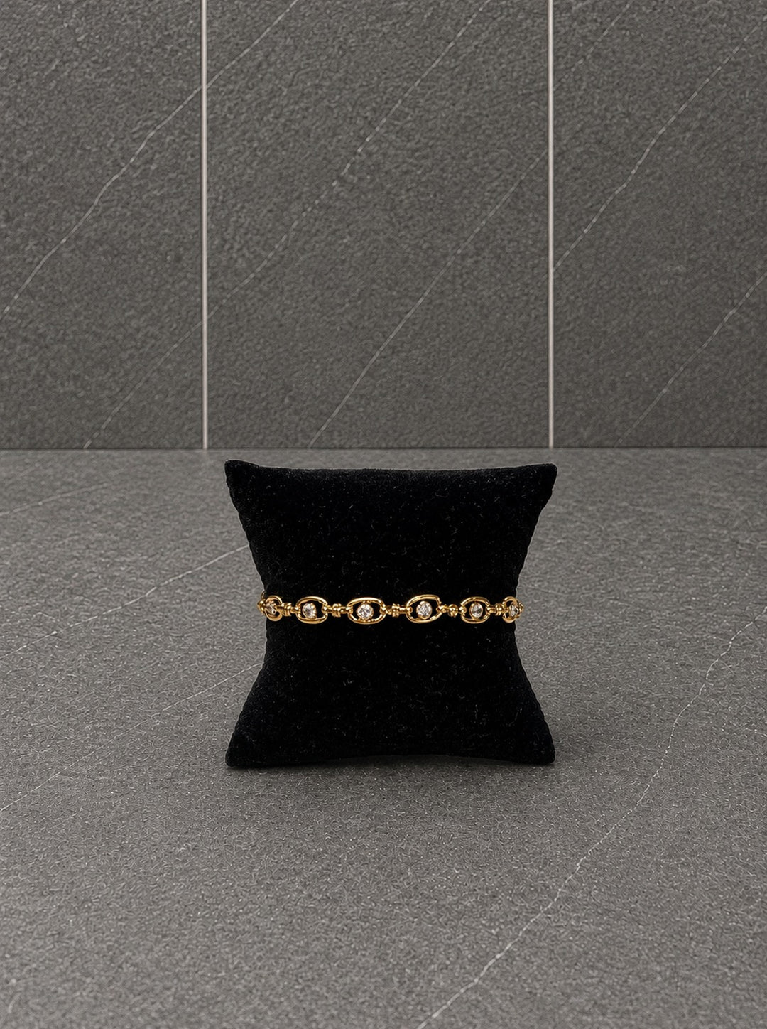 A gold bracelet with oval links and small clear stones, displayed on a black velvet pillow against a gray stone-textured background.