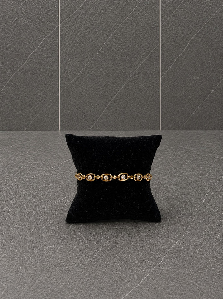 A gold bracelet with oval links and small clear stones, displayed on a black velvet pillow against a gray stone-textured background.