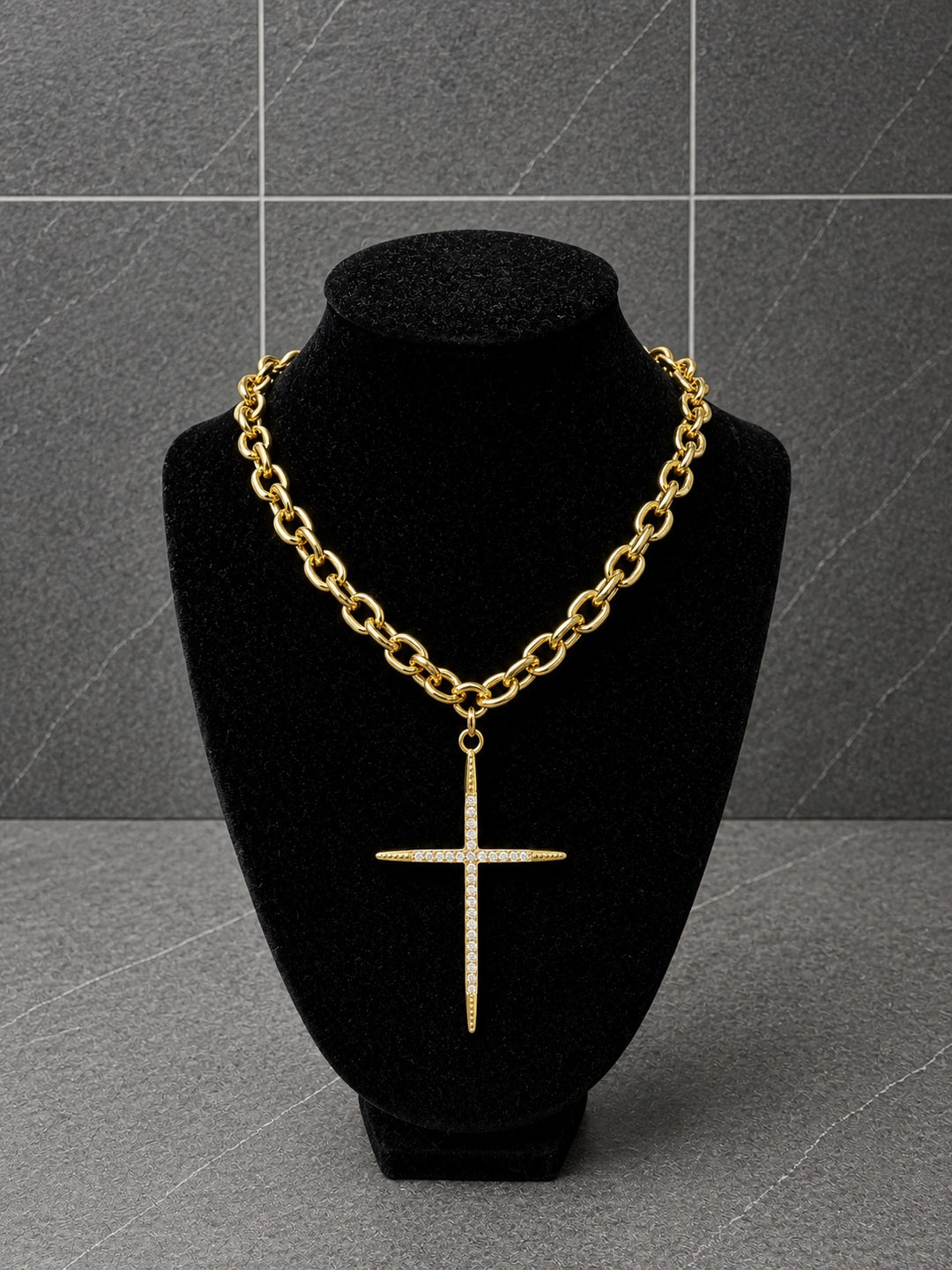 A gold chain necklace with a cross pendant displayed on a black velvet jewelry bust against a gray tiled background.