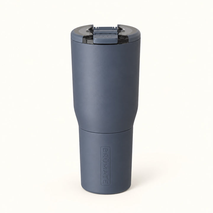 BrüMate Nav 35oz insulated stainless steel travel tumbler in Nightfall Blue, featuring a sleek matte dark blue finish, 100% leakproof BevLock lid with MagFlip technology, a cupholder-friendly base, and a removable color-matched protective sleeve with embossed BrüMate branding. The tall travel coffee tumbler is displayed upright against a soft cream studio background.