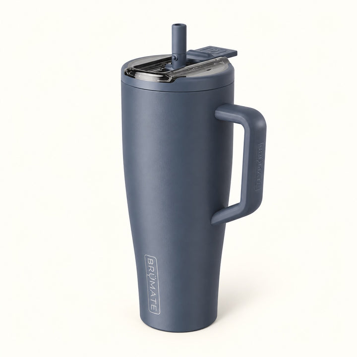 A matte slate blue insulated travel tumbler with a built-in handle, clear lid, and straw, shown on a clean off-white background. The brand name &