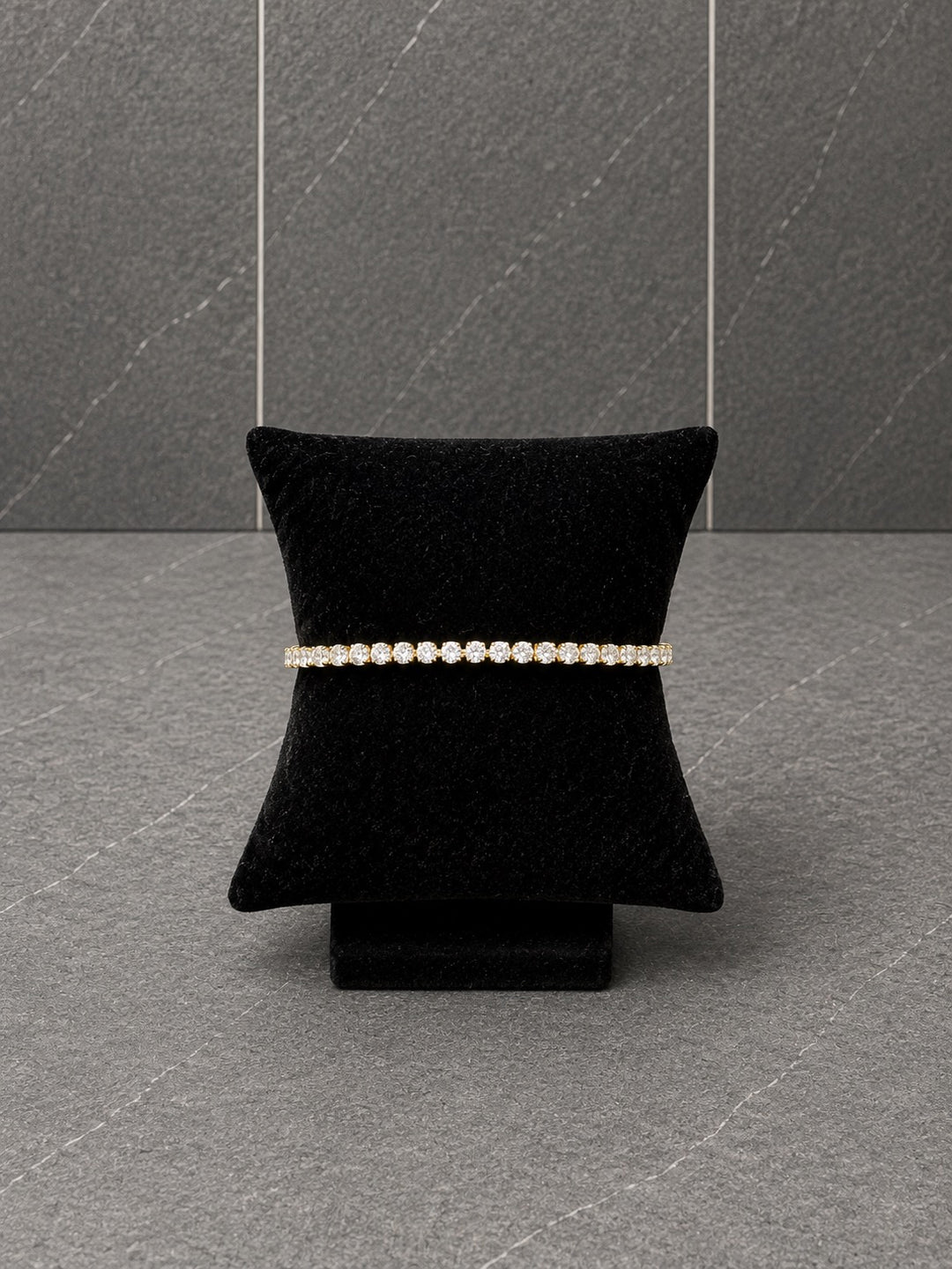 A delicate gold tennis bracelet displayed on a black velvet jewelry stand against a gray textured background.