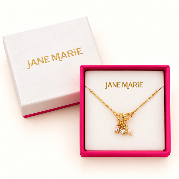 Jane Marie gold pink crystal heart charm necklace displayed in an open pink jewelry box beside its white Jane Marie gift box lid on a cream background.