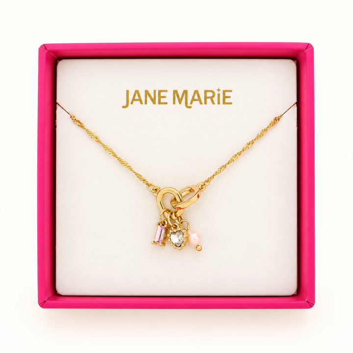 Jane Marie gold chain necklace featuring a pink baguette crystal, sparkling heart-shaped crystal, and pink bead charms displayed in a bright pink jewelry gift box.