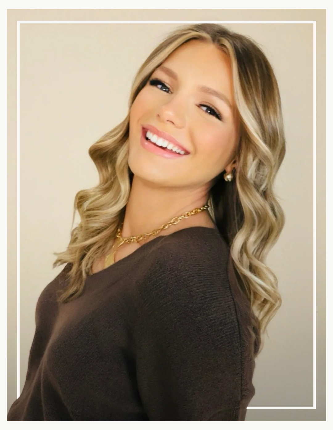 Woman with wavy blonde hair wearing a brown top and gold necklace on a beige background