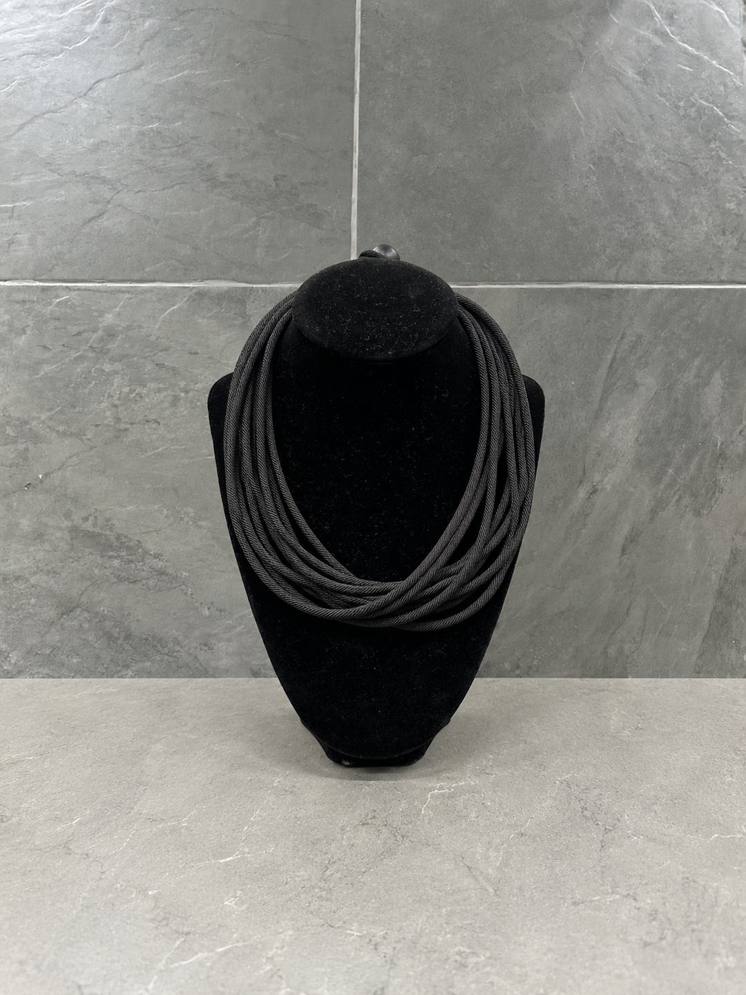 Black multi-strand cord statement necklace displayed on a black velvet jewelry bust with a contemporary gray stone backdrop.