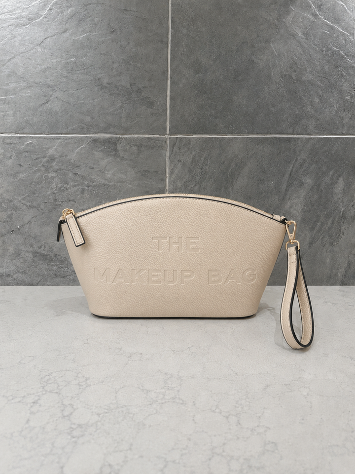 Ivory pebbled faux leather makeup bag with embossed “THE MAKEUP BAG” lettering, gold-tone zipper and detachable wristlet strap, styled on a light marble countertop against a gray stone tile background.