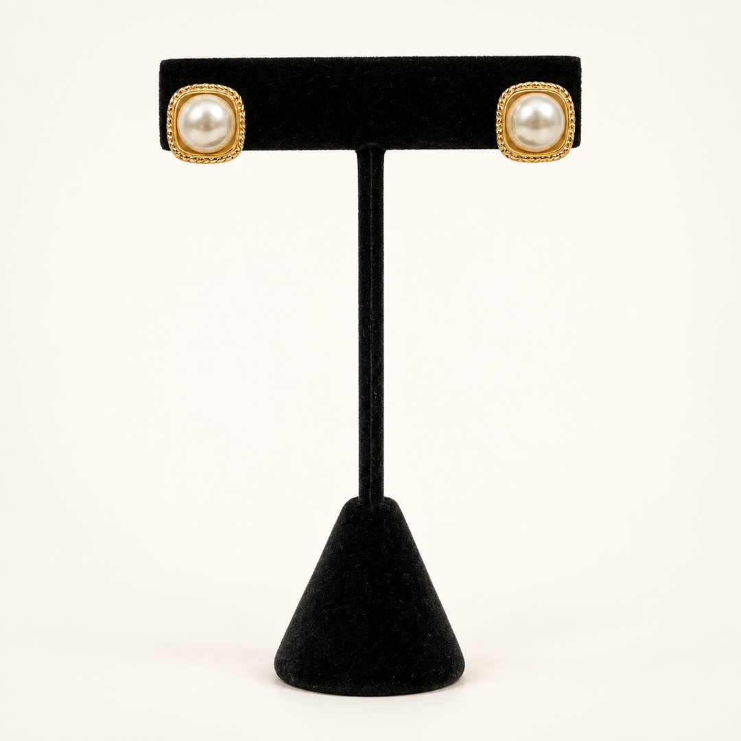 Front view of ivory faux pearl stud earrings with textured gold square frames displayed on a black velvet stand.