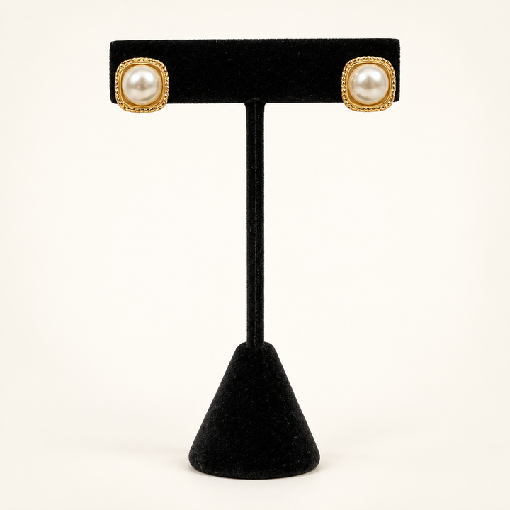 Front view of ivory faux pearl stud earrings with textured gold square frames displayed on a black velvet stand.