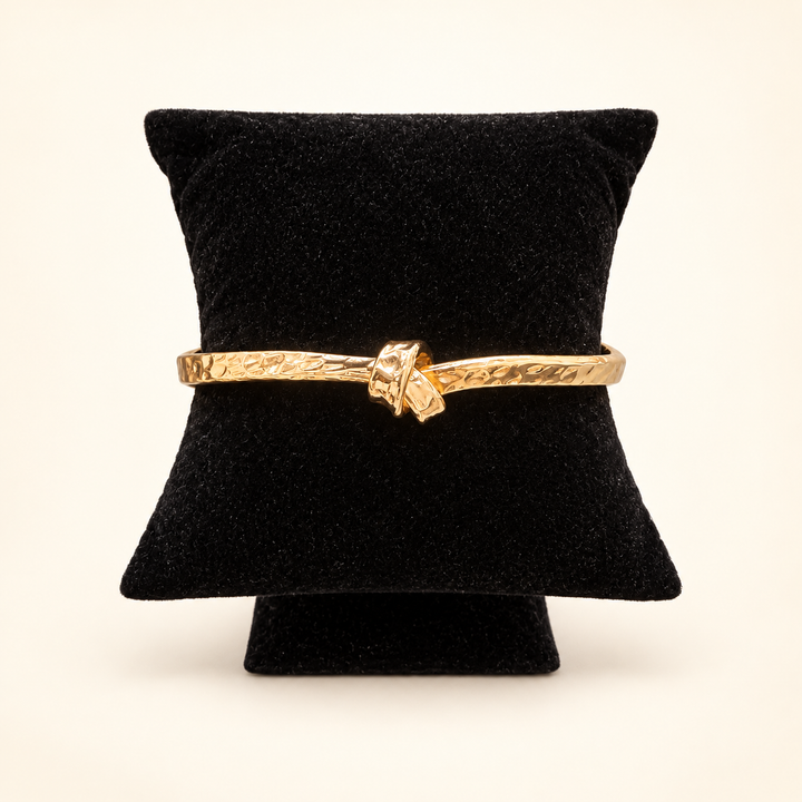 Hammered gold cuff bracelet with a centered knot detail displayed on a black velvet pillow against a cream background.