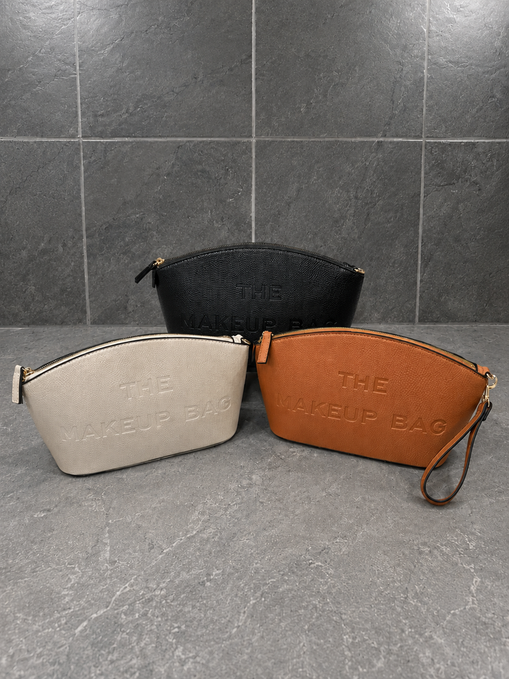 Black, ivory, and camel pebbled faux leather makeup bags with embossed “THE MAKEUP BAG” lettering, gold-tone hardware, and detachable wristlet straps displayed together on a marble countertop against a gray stone tile backdrop.